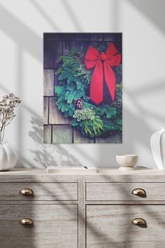 a christmas wreath with a red bow hanging on a wall next to drawers and vases