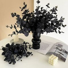 a vase filled with black flowers next to an open book on a white table top