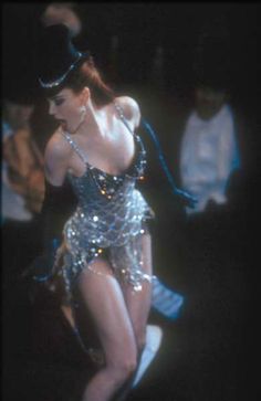 a woman in a silver dress and hat on stage with other people behind her watching