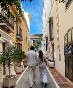 a man and woman walking down an alley way with potted plants on either side