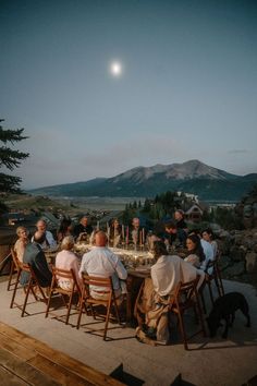 a group of people sitting around a table on top of a wooden deck next to mountains