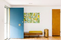 a room with wood floors and white walls, blue door and yellow bench in the middle