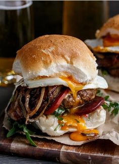 two burgers with meat, cheese and onions on a cutting board next to glasses of beer