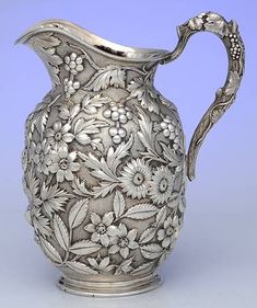 an ornate silver pitcher with flowers and leaves on the handle is shown against a blue background