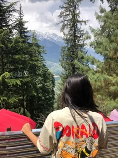 a woman is sitting on a bench looking out at the mountains and trees in the distance