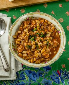 a bowl filled with beans and meat next to a spoon on a green table cloth