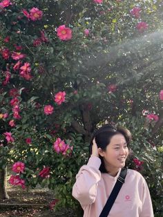 a woman is standing in front of a tree with pink flowers on it and holding her hand up to her ear