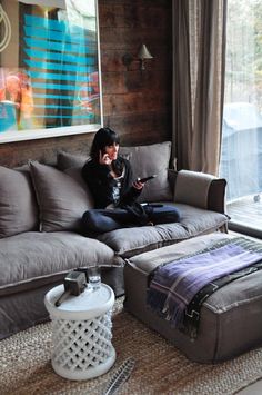 a woman sitting on top of a gray couch in front of a window next to a foot stool