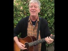 the man is playing his guitar outside in front of some bushes and trees, smiling at the camera