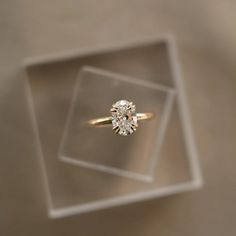 an engagement ring sits in a clear display case on a beige surface with a white background