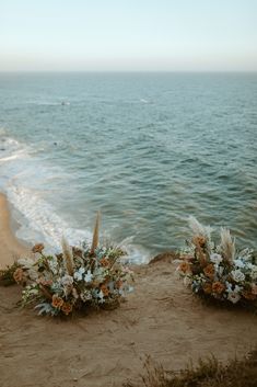 two flower arrangements sitting on top of a sandy beach next to the ocean with waves coming in