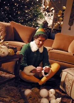 a man sitting on the floor in front of a christmas tree wearing an elf costume