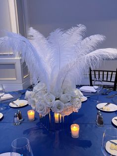 a centerpiece with white flowers and feathers sits on a blue table cloth surrounded by candles