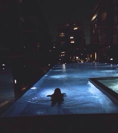 a person swimming in a pool at night with buildings in the backgrouds