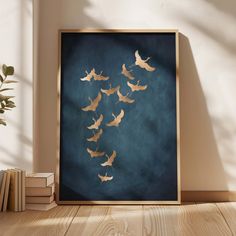 a framed photograph of birds flying in the air on a wooden floor next to books and a potted plant