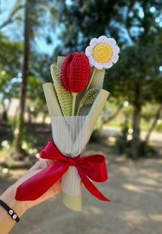 a hand holding a bouquet of flowers with a red ribbon tied around the stems and two crocheted flower heads