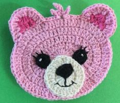 a crocheted pink bear with black eyes on a green background, made to look like it is smiling