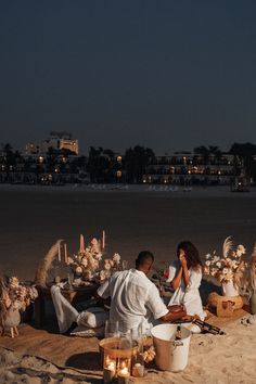 a man and woman sitting on the beach next to each other with candles in front of them