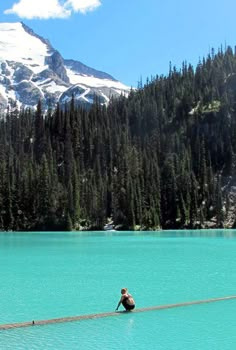 a person sitting on a log in the middle of a lake with mountains in the background