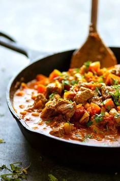 a skillet filled with meat and vegetables on top of a wooden table next to a spoon