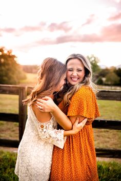 two women hugging each other in front of a fence and grass field with the sun setting behind them