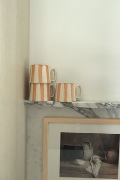 two coffee mugs sitting on top of a marble shelf next to a framed photograph