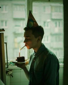 a man with a birthday hat blowing out a candle on a cupcake in front of a window