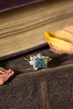 The "Aurae" engagement ring from Staghead Designs sits on a stack of vintage books. This 14K yellow gold ring features an east-west set emerald cut moss agate center stone & asymmetrical floral diamond accents. Whiskey Barrel Wedding Ring, Antler Wedding Rings, Agate Wedding Ring, Rings Sets, Agate Wedding, Handcrafted Engagement Ring, Wood Wedding Ring, Moss Agate Engagement Ring, Yellow Gold Engagement Ring