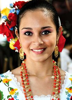 a woman in a colorful dress with necklaces and flowers on her head smiling at the camera