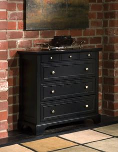 a black chest of drawers in front of a brick wall with a painting on it