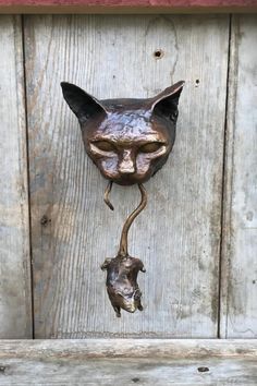 a metal cat head mounted to the side of a wooden door with a rat hanging from it's tail