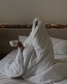 a person wrapped in a blanket sitting on a bed with a coffee cup under their arm