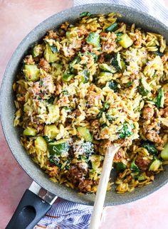 a skillet filled with pasta and vegetables on top of a pink table cloth next to a wooden spoon