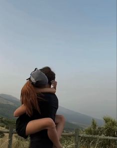 two people sitting on top of each other in front of a fence with mountains in the background