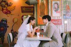 a man and woman sitting at a table in front of coca - cola machines with signs on the wall behind them