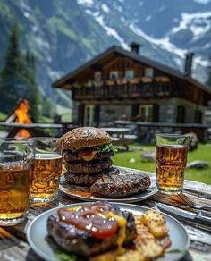 two hamburgers and some drinks on a wooden table in front of a mountain cabin