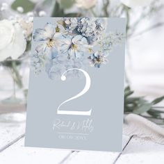 a wedding table number with blue flowers and greenery in the background, on a white wooden surface