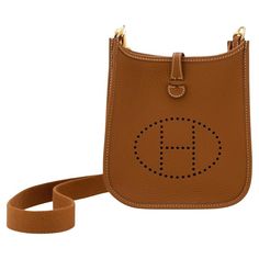 Hermès mini Evelyne in gold Clemence leather complemented with gold-tone hardware and white stitching. It features a perforated "H" design, a removable shoulder strap, an open interior, and a snap lock closure. Date stamp is W 2024 The dimensions are 16cm L x 20cm H x 5cm D. The bag comes complete with a Hermès box, dust bag, removable strap, care book. Hermes Small Crossbody Bag, Hermes Crossbody Bag, Snap Lock, Hermes Box, Fashion Handbags, Purses Crossbody, Gold Hardware, Messenger Bag, 20 Cm