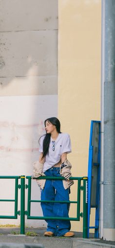 a woman standing on the side of a road next to a building with a green fence