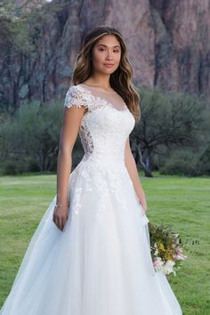 a woman in a white wedding dress standing on grass with mountains in the back ground