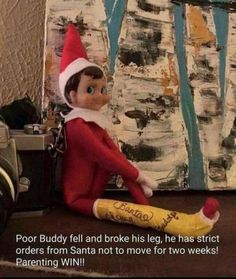 an elf sitting on top of a table next to a painting with the caption poor buddy fell and broke his leg, he has strict orders from santa not to move for two weeks