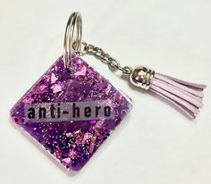 a purple glitter square keychain with an antihero message on it and tassels