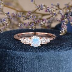 a white opal and diamond ring sitting on top of a blue velvet cushion with purple flowers in the background