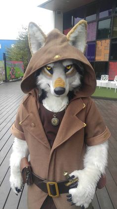 a stuffed animal dressed up like a fox wearing a trench coat and hat, standing on a wooden deck