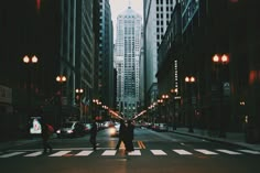 two people are crossing the street in front of tall buildings at night with lights on