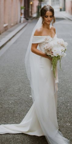 the bride is walking down the street with her bouquet in hand and she is wearing an off shoulder wedding dress