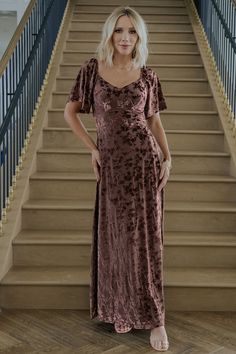 a woman standing in front of stairs wearing a brown velvet dress with ruffle sleeves