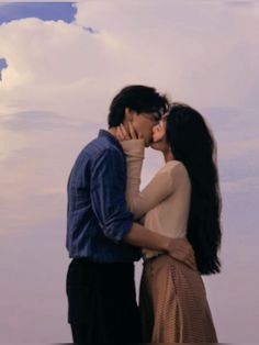 a man and woman kissing each other in front of a sky with clouds behind them