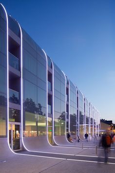 people are walking around in front of a large building with curved glass walls at dusk