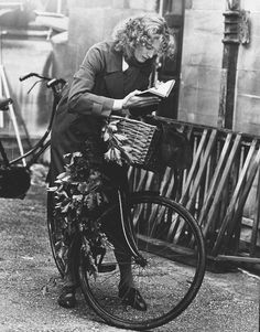 an old black and white photo of a woman on a bicycle reading a book while holding a basket full of flowers